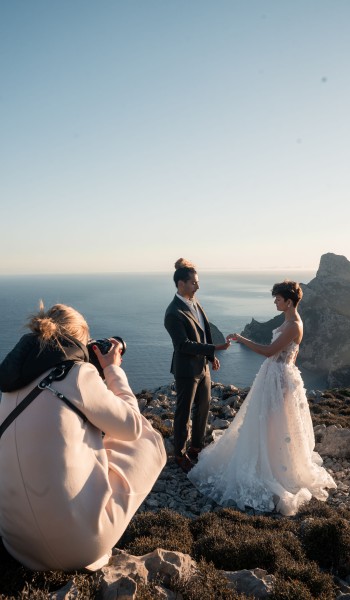 Fotograf kniet, fotografiert Brautpaar auf Felsen mit Meerblick. Braut im weißen Kleid, Bräutigam im Anzug.