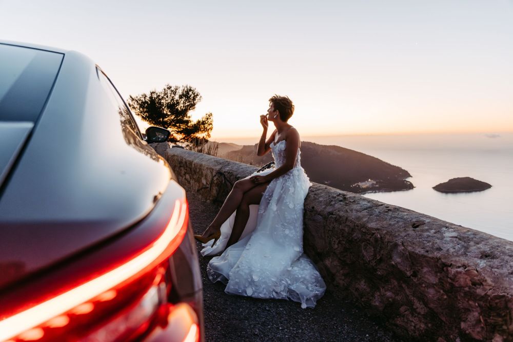 Frau im Brautkleid sitzt bei Sonnenuntergang auf einer Mauer, neben einem Auto, mit Blick auf das Meer und Hügel.