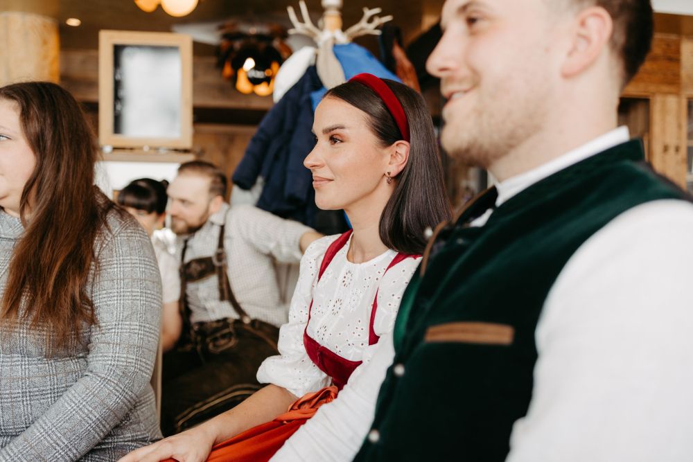 Menschen in traditioneller Kleidung sitzen in einem Raum. Eine Frau trägt ein rotes Haarband und eine weiße Bluse.