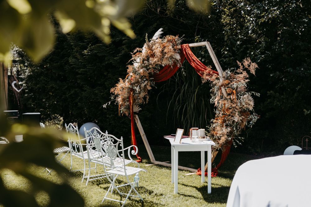 Ein Hochzeitsbogen mit roten Tüchern und Trockenblumen, daneben weiße Bänke und ein Tisch im Garten.