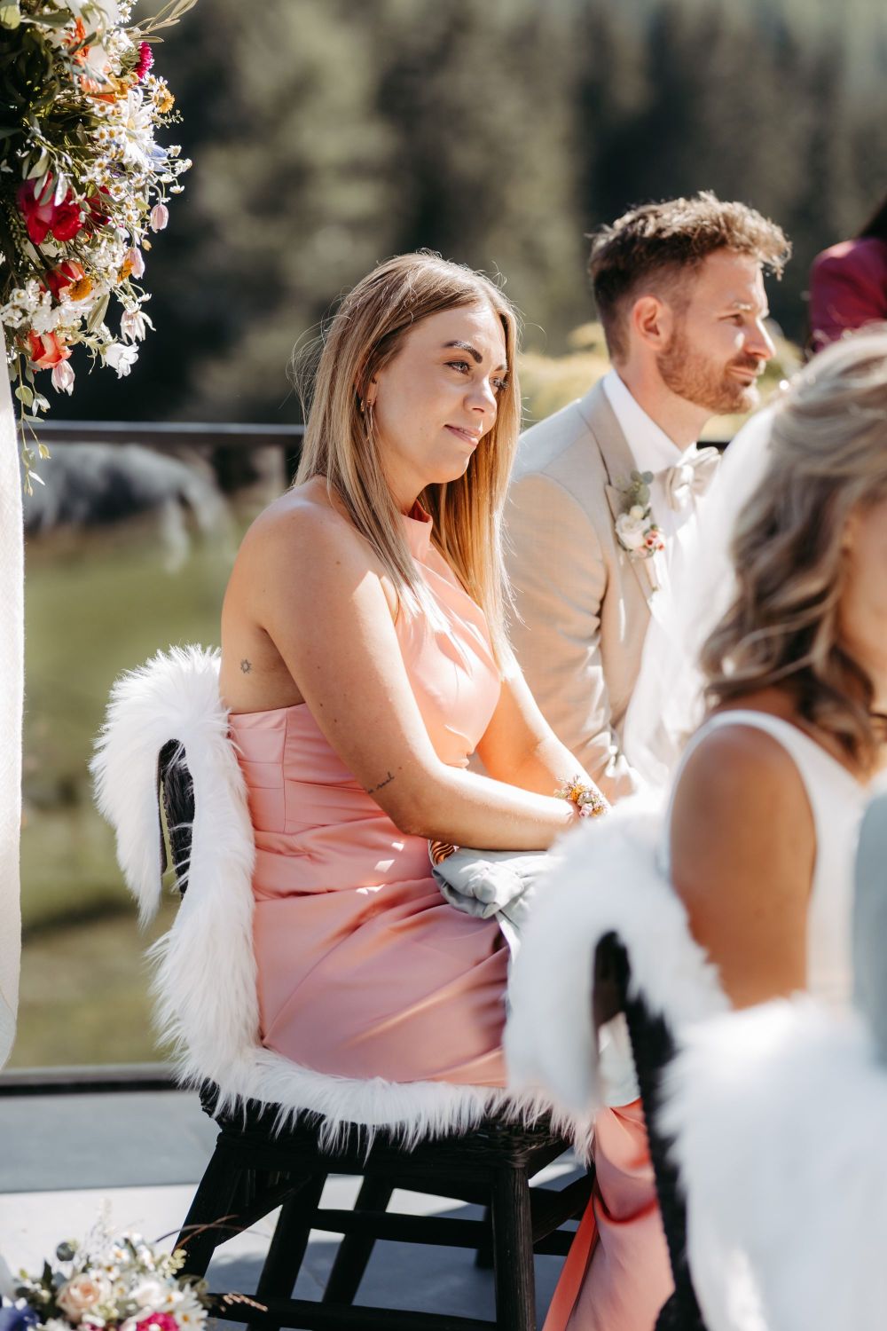 Frau in rosa Kleid sitzt draußen, wirkt nachdenklich und ruhig. Im Hintergrund Natur und Blumenarrangement.
