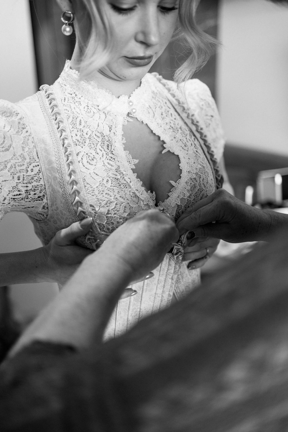 Person in einem Spitzenkleid wird von einer anderen Person beim Schließen der Knöpfe an der Vorderseite des Kleides unterstützt.