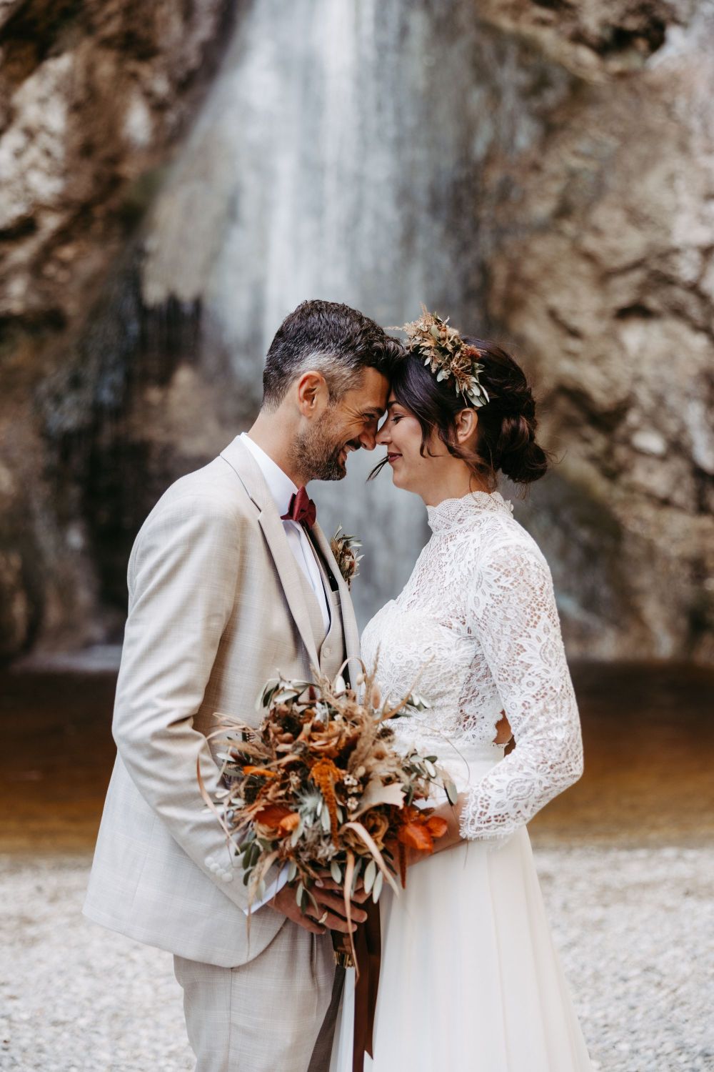 Ein Paar in Hochzeitskleidung steht lächelnd Stirn an Stirn vor einem Wasserfall, die Braut hält einen Blumenstrauß.