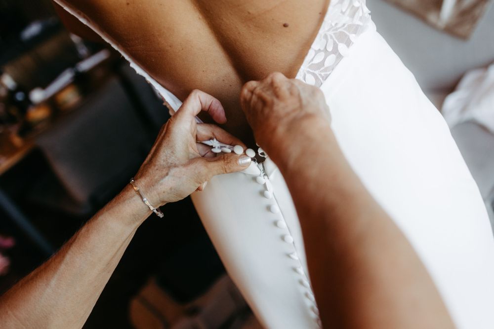 Nahaufnahme einer Hand, die Knöpfe am Rücken eines eleganten weißen Kleides schließt. Symbol für Hingabe und Detail.
