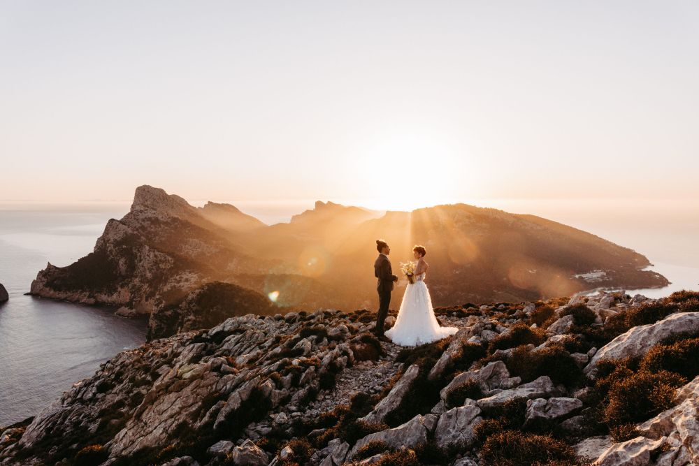 Ein Paar steht in Hochzeitskleidung auf einem felsigen Berg, im Hintergrund Sonnenuntergang über dem Meer.