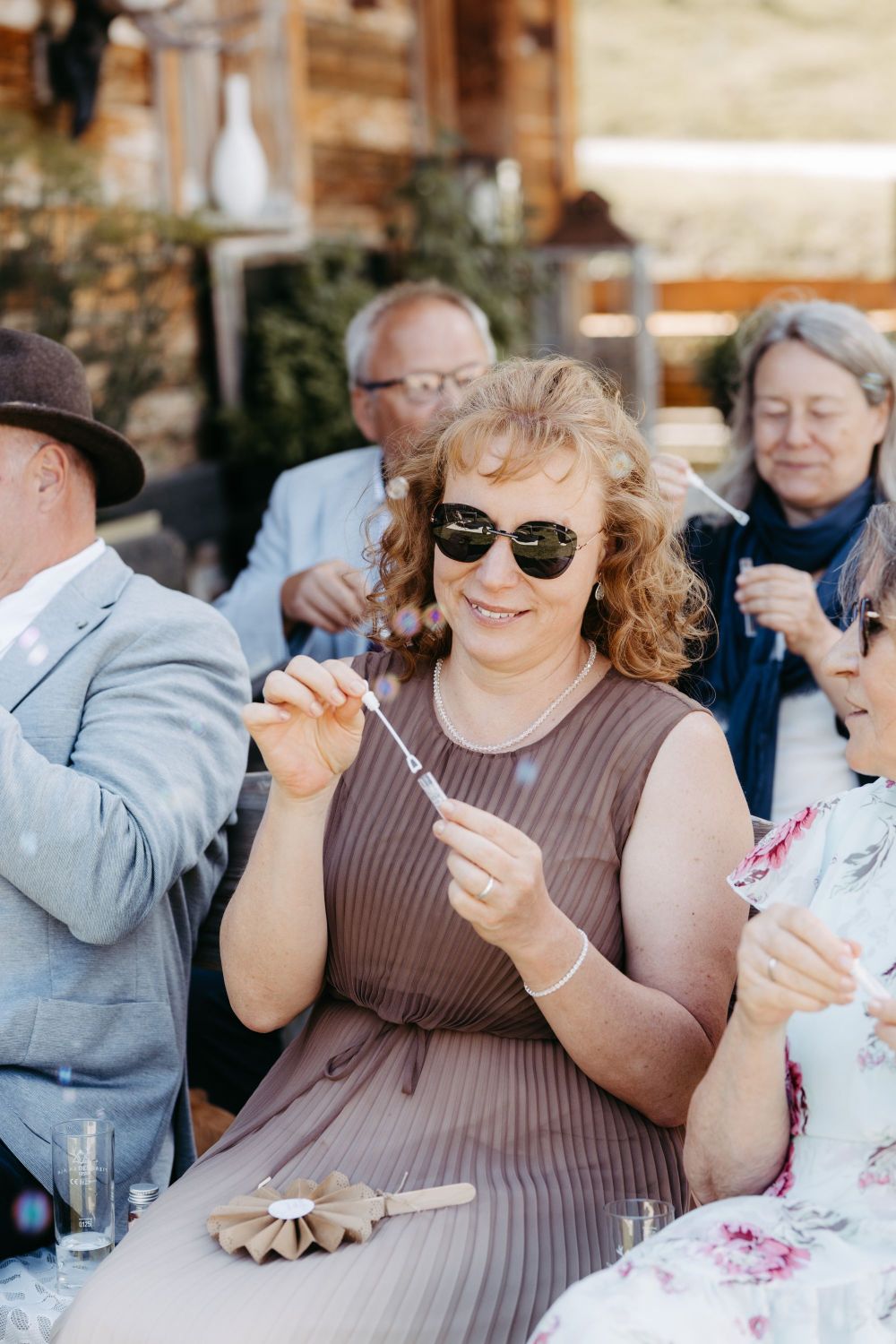 Frau in Sonnenbrille lächelt und bläst Seifenblasen in einer geselligen Runde im Freien – Berghochzeit auf der Tiergartenalm, Mühlbach