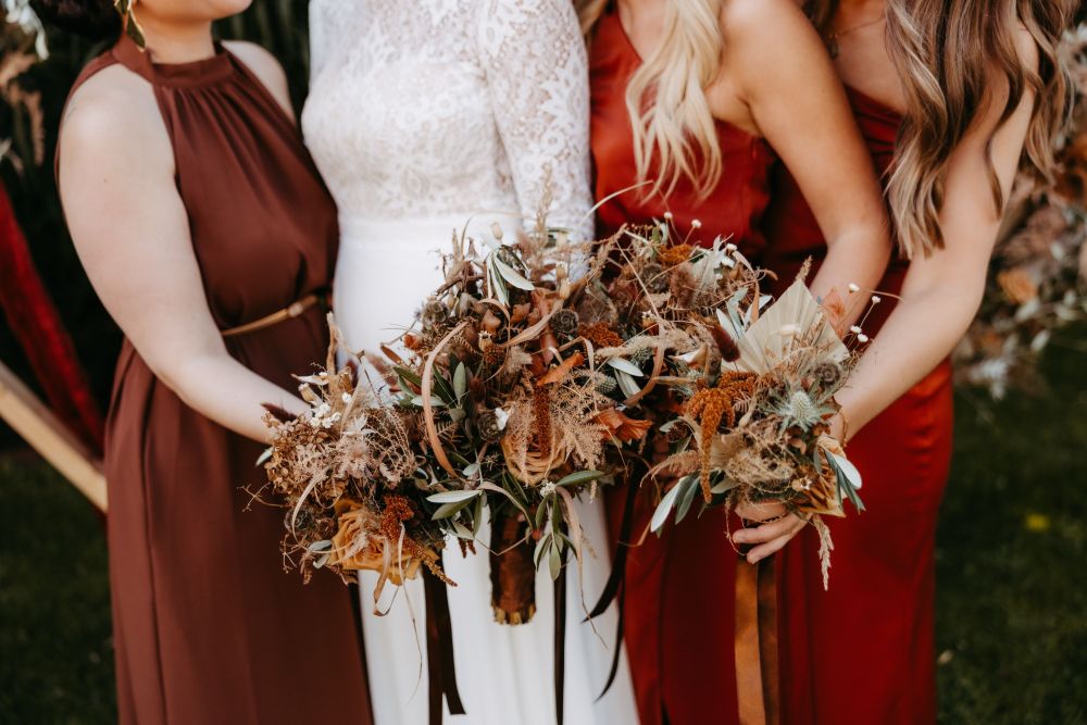 Drei Frauen in eleganten Kleidern halten herbstliche Blumensträuße mit braunen und grünen Tönen.