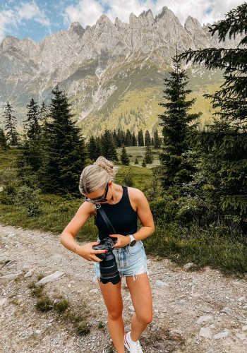 Person mit Kamera auf einem Wanderweg vor einer Berglandschaft mit Tannen und felsigen Gipfeln.