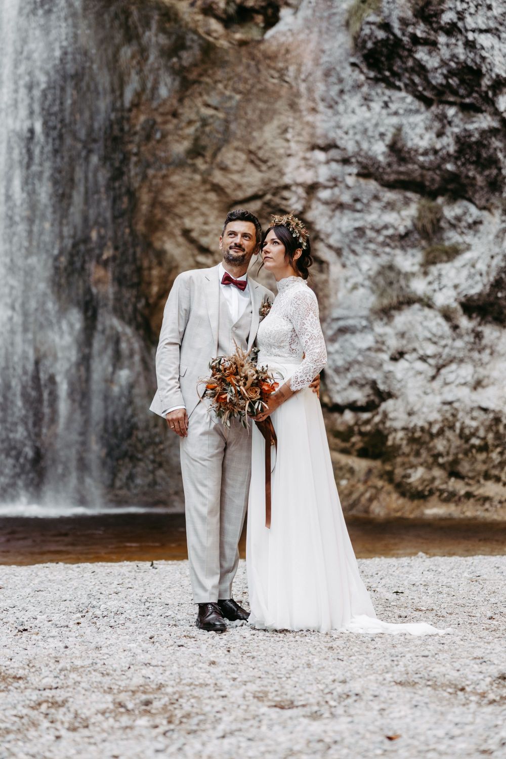 Ein Paar in eleganter Hochzeitskleidung steht vor einem Wasserfall auf Kiesboden. Die Frau hält einen Blumenstrauß.