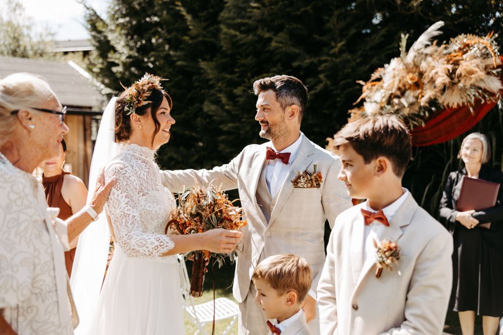 Braut und Bräutigam lächeln sich bei einer Outdoor-Hochzeit an. Zwei Jungen und eine ältere Frau sind ebenfalls im Bild.