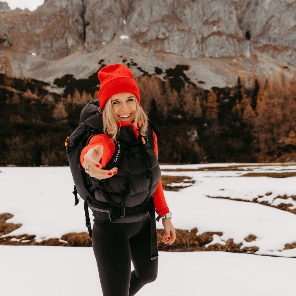 Frau in rotem Hut und schwarzer Jacke steht lächelnd im Schnee, reicht die Hand aus. Berglandschaft im Hintergrund.