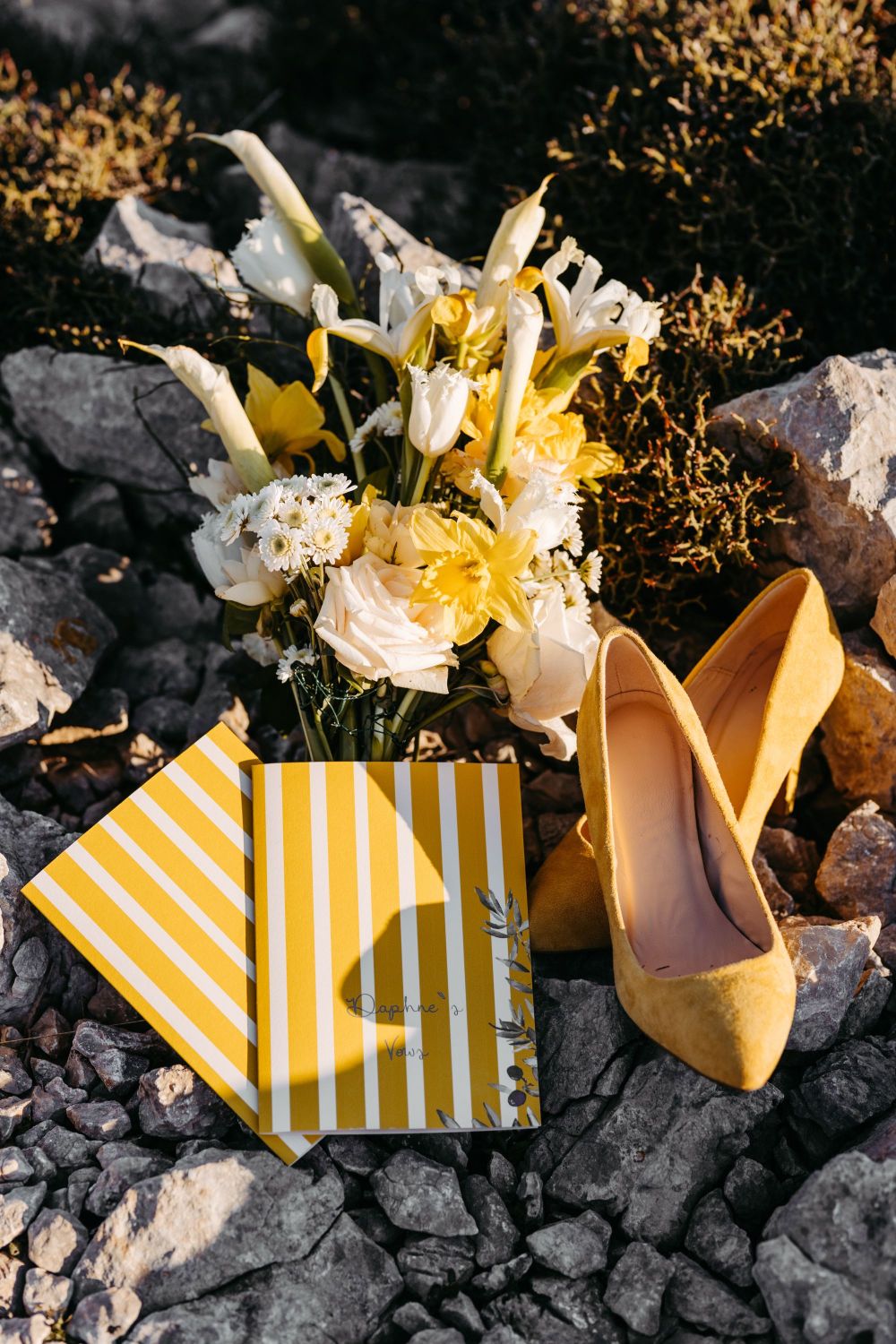 Gelbe Schuhe, ein Blumenstrauß mit gelben und weißen Blumen und ein gelb-weiß gestreiftes Heft auf grauen Steinen.
