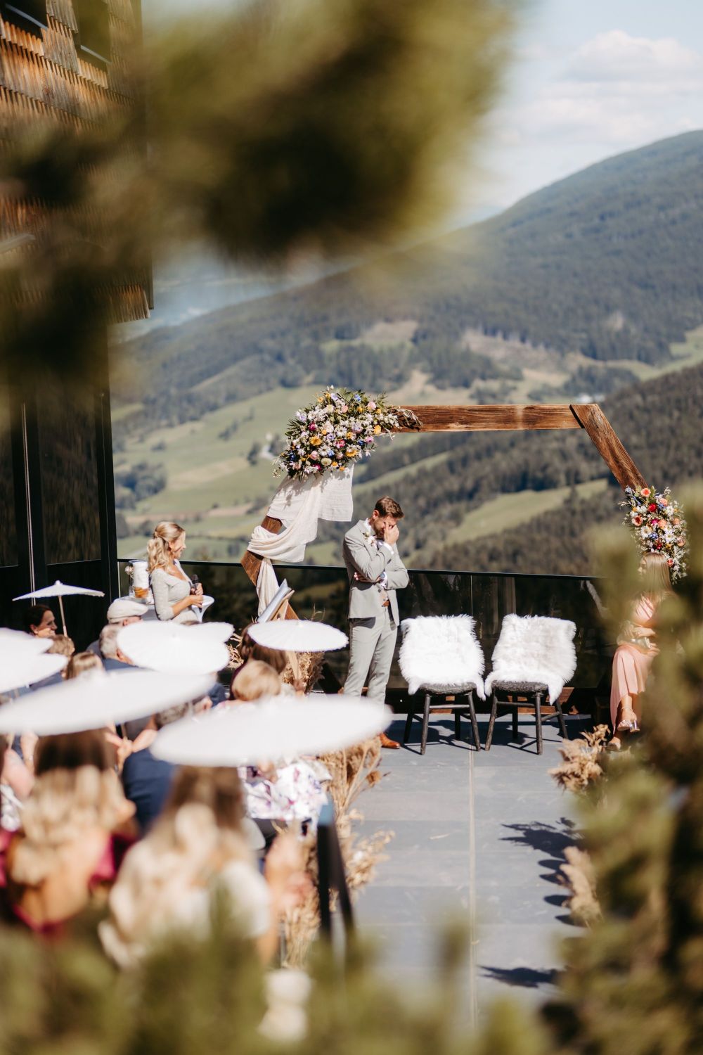 Outdoor-Zeremonie mit Bergblick, Blumenbogen und Gästen mit weißen Sonnenschirmen. Ein Mann im Anzug wirkt emotional.