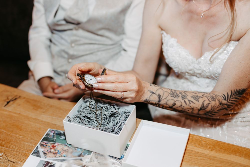 Person in Hochzeitskleid hält eine Taschenuhr über einer Geschenkbox. Symbol für Zeit und Lebensentscheidungen – Berghochzeit auf der Tiergartenalm, Mühlbach