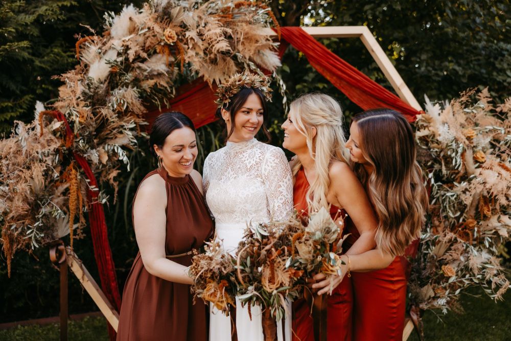 Vier Frauen in eleganten Kleidern lachen vor einem Blumenbogen im Freien. Eine trägt ein weißes Spitzenkleid.