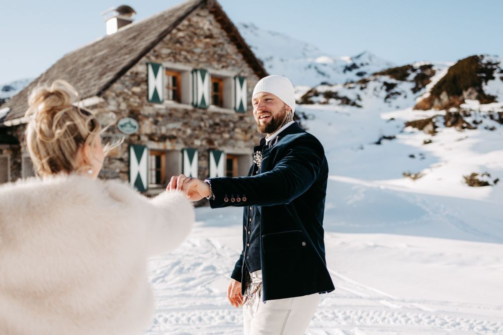 Ein Mann und eine Frau tanzen lachend im Schnee vor einer Berghütte, umgeben von verschneiten Bergen.