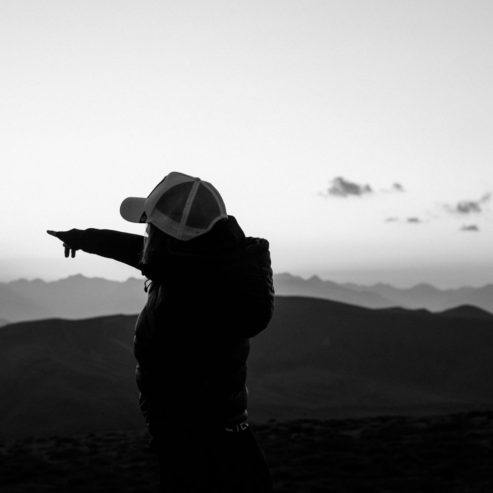 Person mit Baseballkappe zeigt in die Ferne. Silhouette vor Berglandschaft bei Sonnenuntergang. Schwarz-Weiß-Foto.