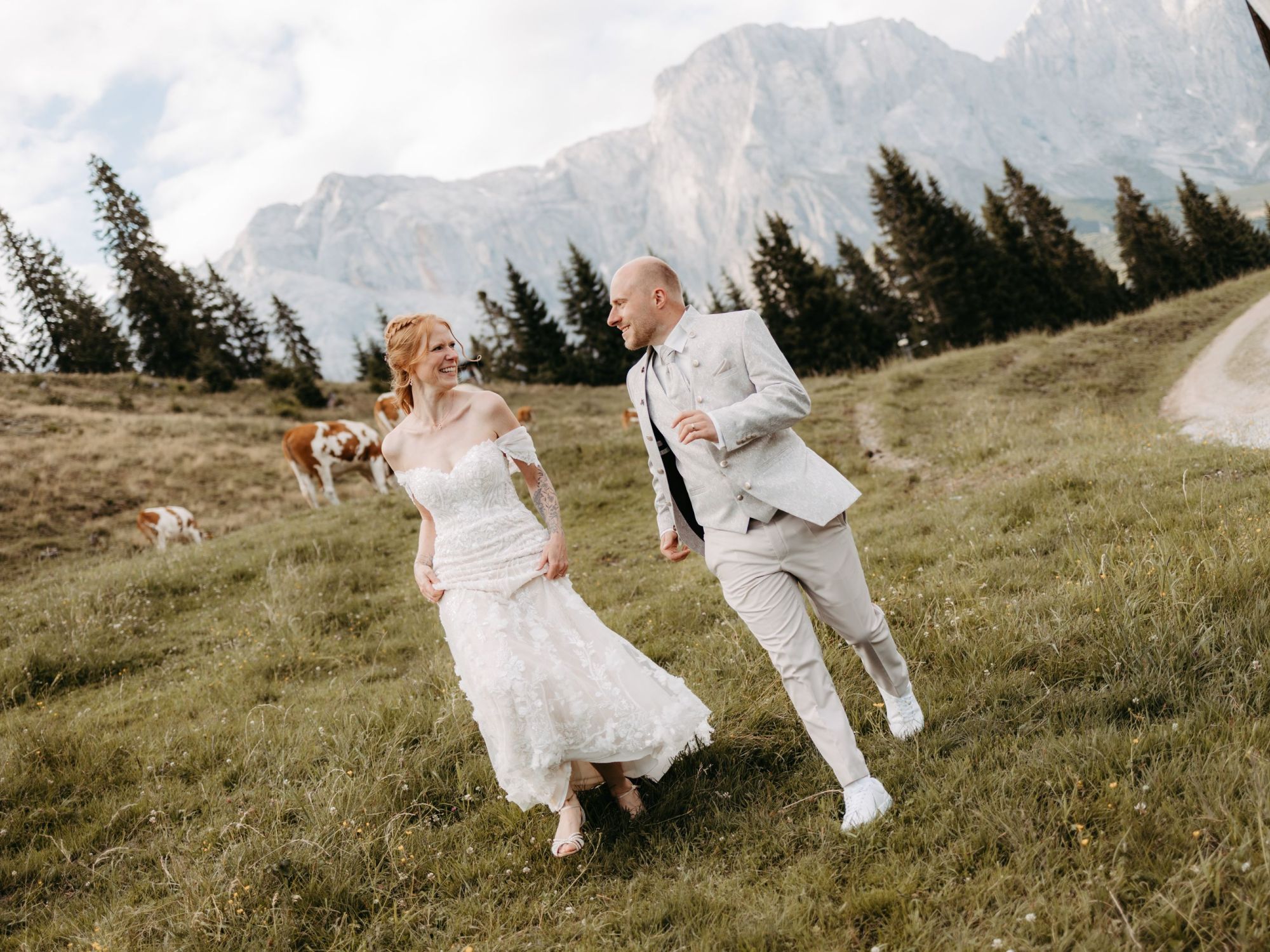 Paar in eleganter Kleidung läuft lachend über eine Wiese vor Bergkulisse, symbolisiert Lebensfreude und neue Wege – Berghochzeit auf der Tiergartenalm, Mühlbach