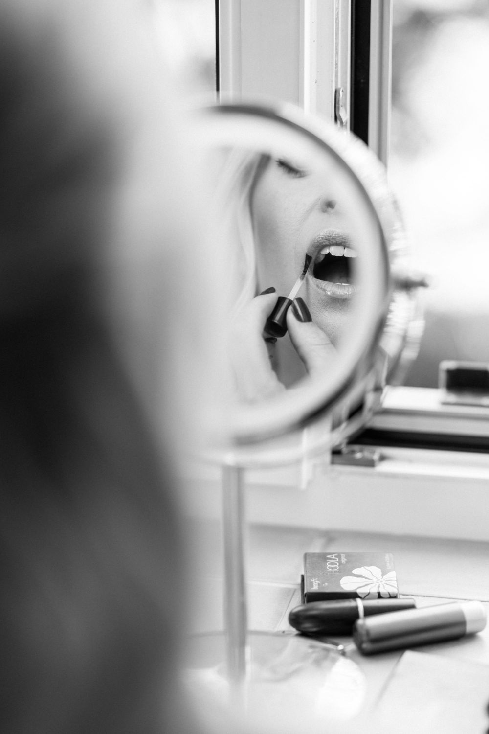 Person trägt Lippenstift auf, im Spiegel sichtbar. Schminkutensilien liegen auf dem Tisch. Schwarz-weiß Fotografie.