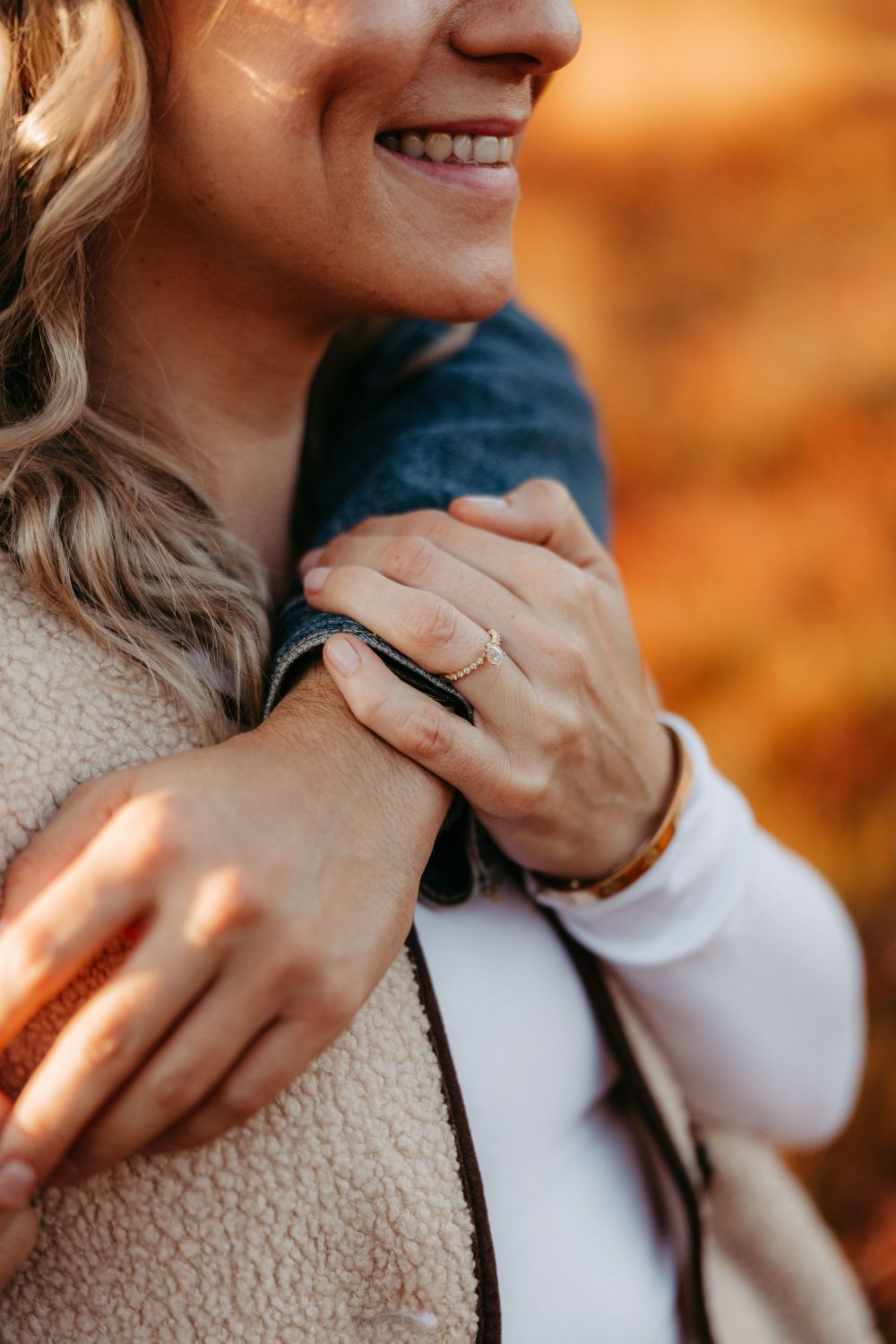 Nahaufnahme einer lächelnden Frau, die eine Hand mit Ring über eine andere legt, vor herbstlichem Hintergrund.
