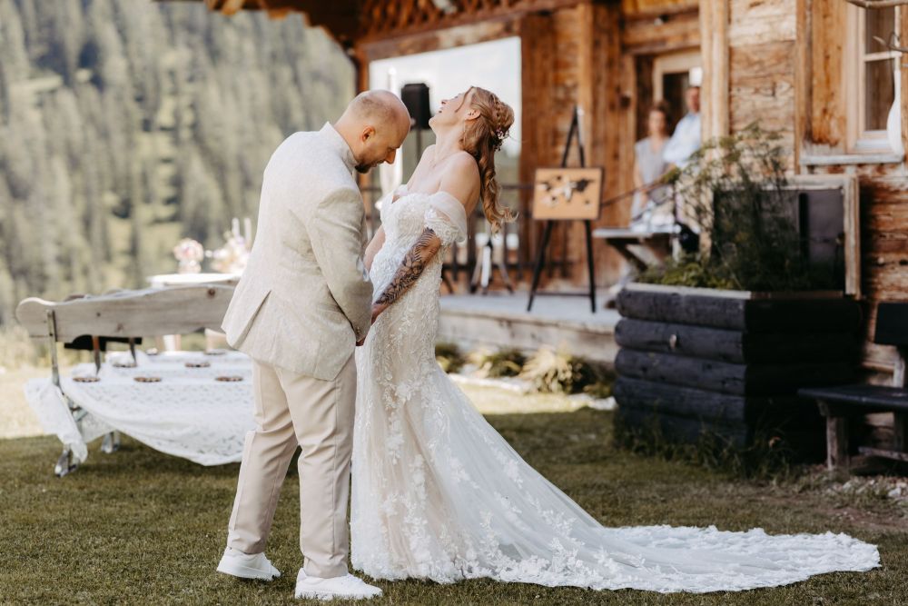 Ein Paar tanzt lachend in Hochzeitskleidung vor einer rustikalen Hütte in den Bergen, umgeben von Natur – Berghochzeit auf der Tiergartenalm, Mühlbach