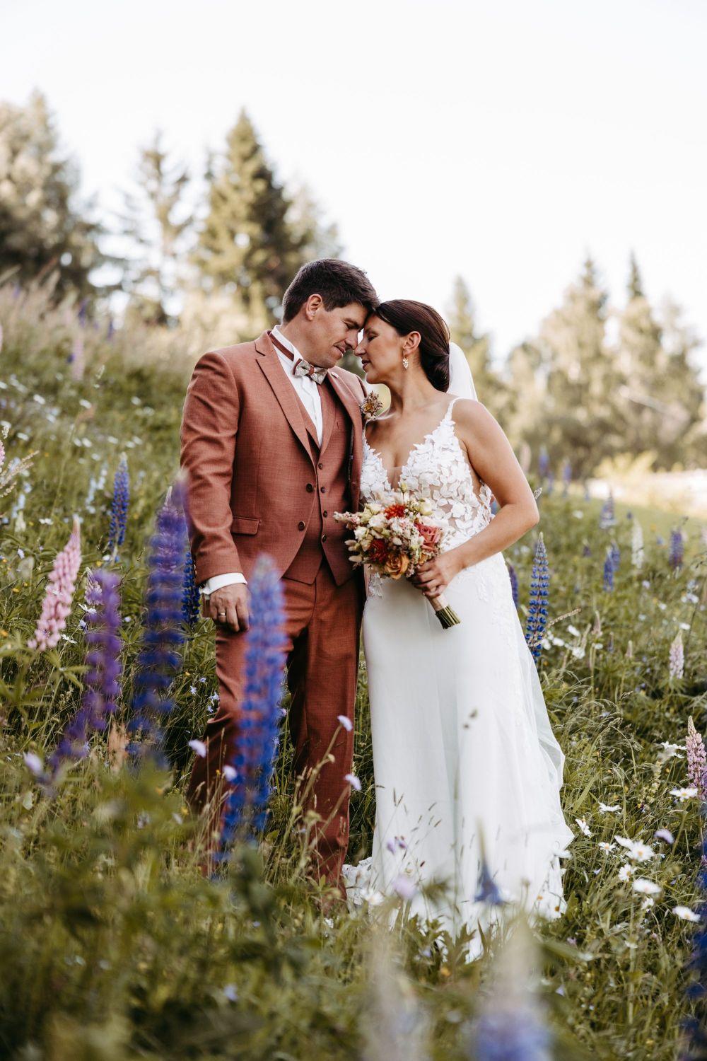 Ein Paar in Hochzeitskleidung steht in einer blühenden Wiese, Stirn an Stirn, umgeben von Bäumen.