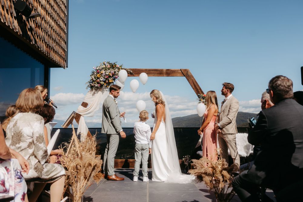 Freiluft-Hochzeit mit Paar und Kind unter Blumenbogen, Gäste im Halbkreis, Fokus auf Lebensfreude und neue Anfänge.