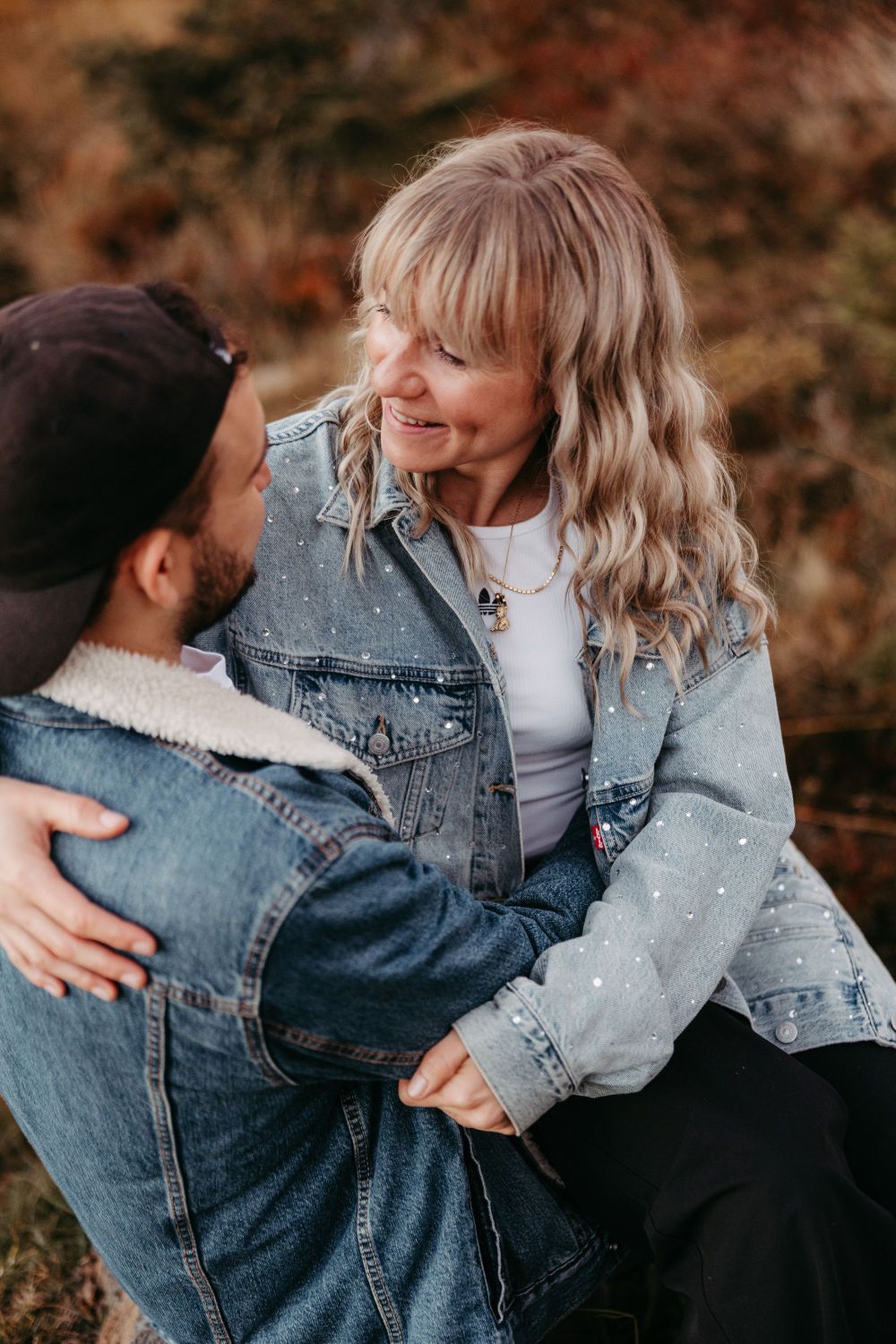Zwei Personen in Jeansjacken umarmen sich und lächeln einander an, in einer herbstlichen Landschaft.