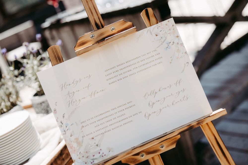 Ein Schild auf einer Staffelei mit der Aufschrift: "Handys aus. Augen auf. Herzen öffnen." im floralen Design – Berghochzeit auf der Tiergartenalm, Mühlbach