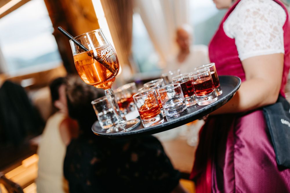 Kellnerin in traditioneller Kleidung serviert Aperitif und Shots auf Tablett in geselliger Runde, symbolisiert Genussmomente – Berghochzeit auf der Tiergartenalm, Mühlbach