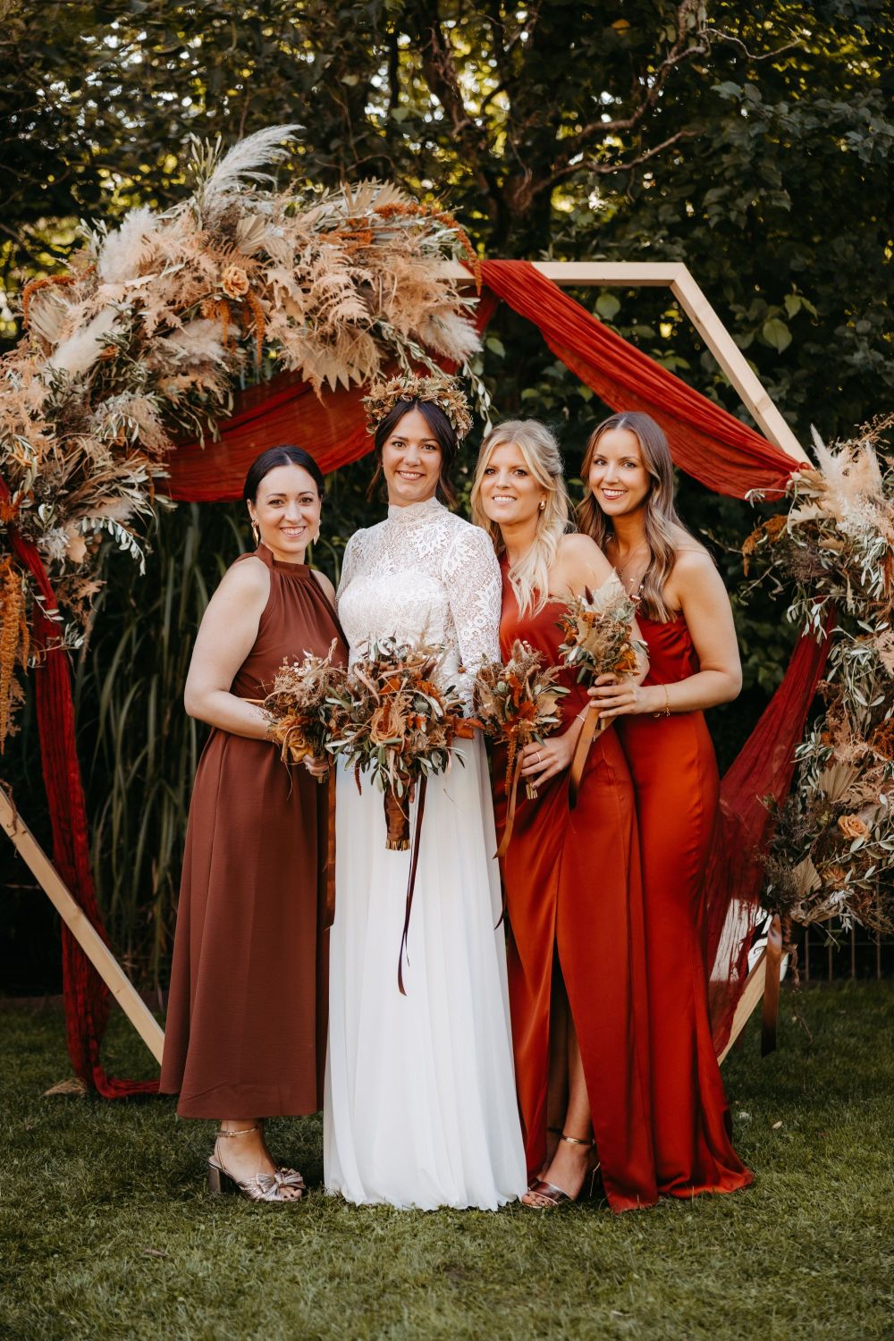 Vier Frauen in eleganter Kleidung posieren vor einem Blumenbogen im Freien. Die Braut trägt ein weißes Kleid.