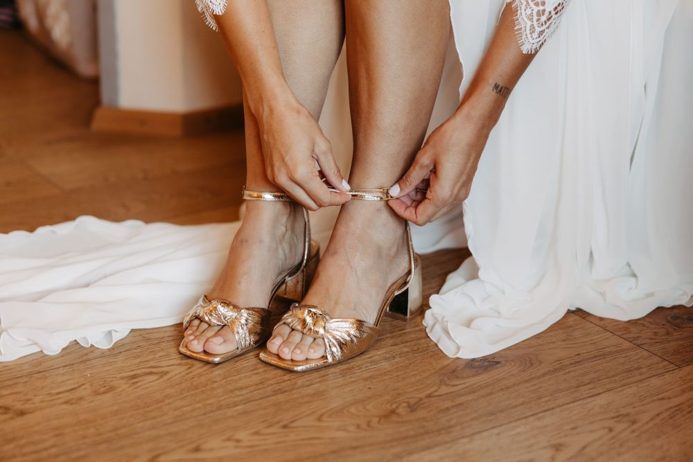 Frau in weißem Kleid schnallt goldene Sandaletten an, sitzt auf Holzboden. Kleid fällt locker auf den Boden.