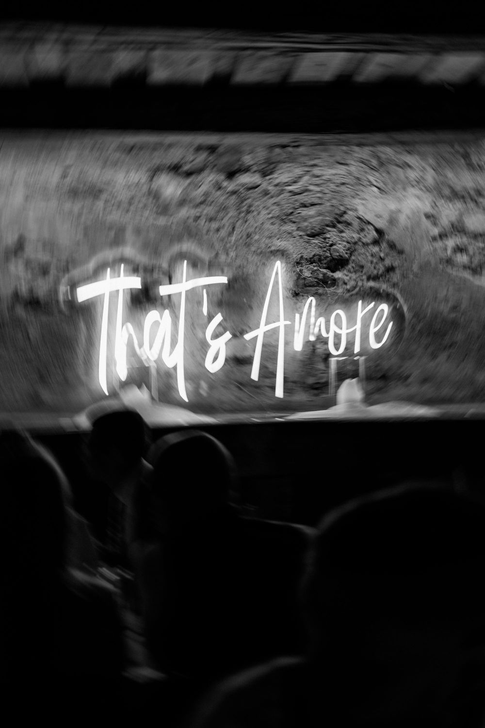 Neon-Schriftzug "That's Amore" vor unscharfem Hintergrund, darunter Silhouetten von Personen.