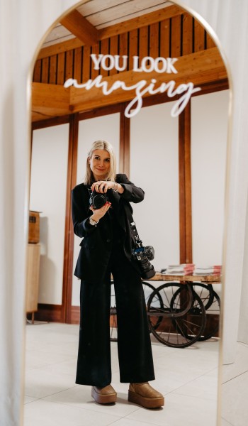 Frau in schwarzem Anzug fotografiert sich im Spiegel mit "You look amazing"-Schriftzug, Kamera in der Hand.