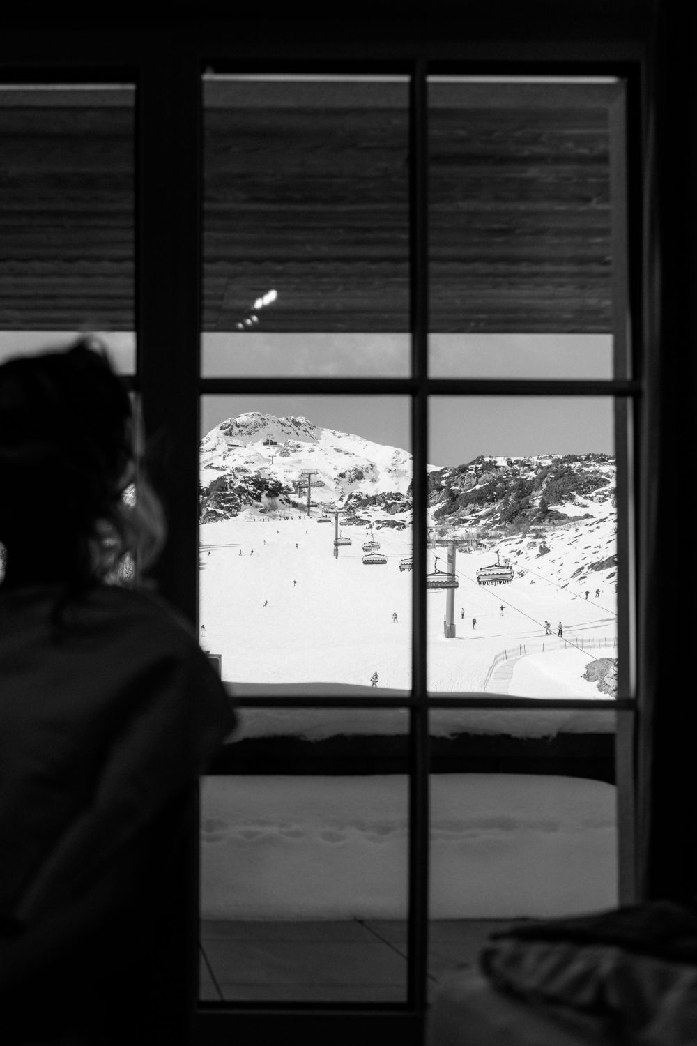 Blick aus einem Fenster auf eine verschneite Berglandschaft mit Skilift und Skifahrern, Person im Vordergrund unscharf.