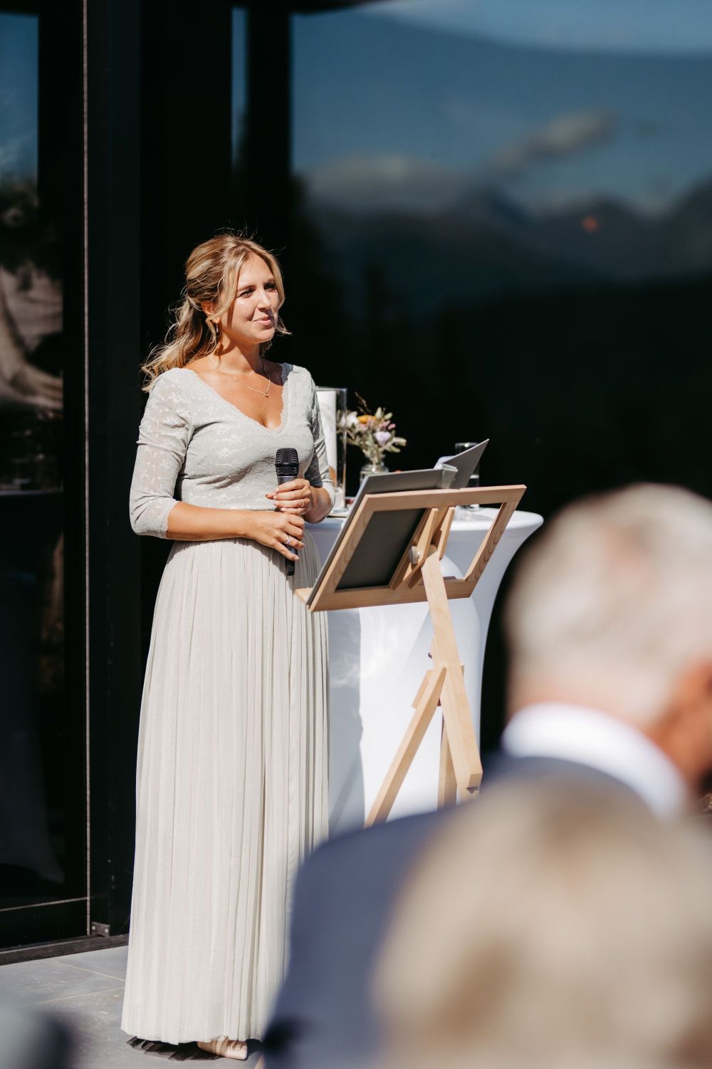 Eine Frau in einem eleganten Kleid spricht draußen mit einem Mikrofon, neben einem Rednerpult.