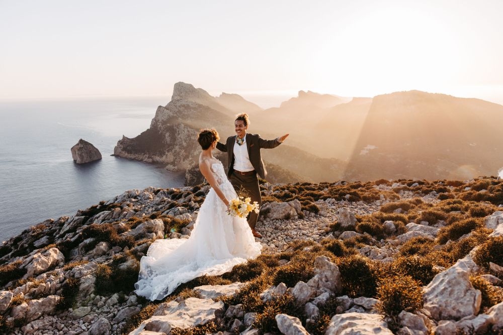Brautpaar in Hochzeitskleidung steht auf felsigem Berggipfel bei Sonnenuntergang, mit Meer und Bergen im Hintergrund.