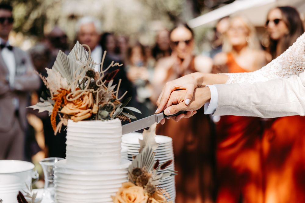 Hände schneiden Hochzeitstorte mit Blumenverzierung, umgeben von Menschen in festlicher Kleidung im Hintergrund.