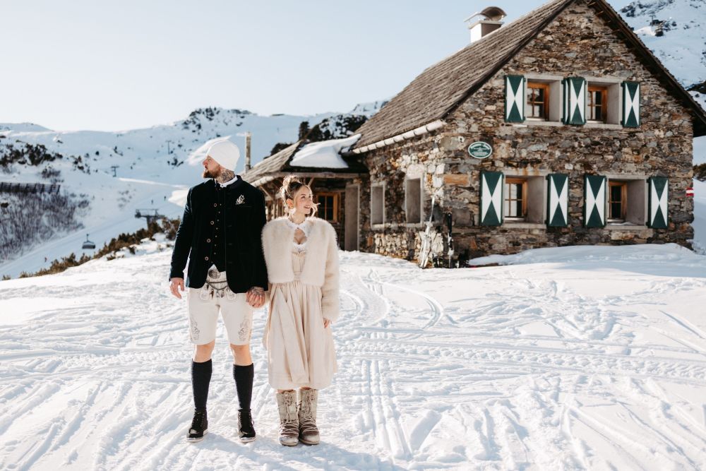Ein Paar in traditioneller Kleidung steht Hand in Hand vor einer verschneiten Berghütte.