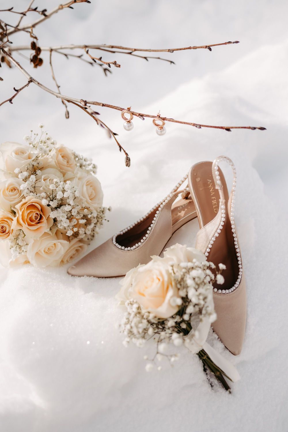 Beige High Heels mit Perlen, Rosenstrauß und Zweig mit Ohrringen im Schnee arrangiert.